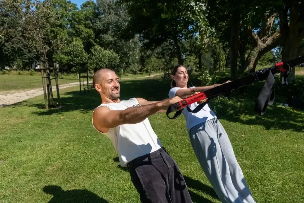 Florian Pirès se tient au premier plan, souriant, aux côtés d'une autre personne qui s'exerce en extérieur avec des bandes de résistance, le tout se déroulant dans un parc.
