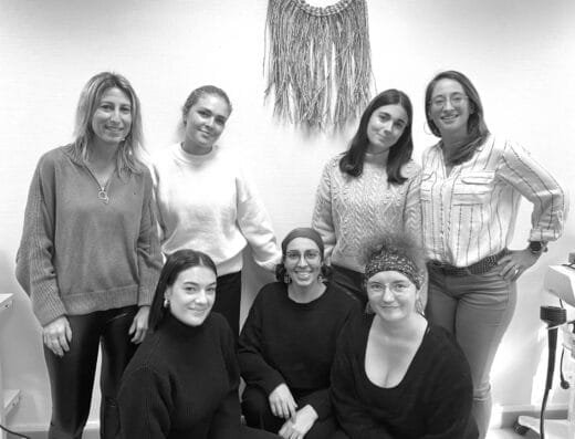 Sept personnes, dont Astrid Chatain, posent et sourient en groupe dans un intérieur. Certaines sont debout, d'autres assises, en tenue décontractée. Une tenture tissée décore le mur derrière elles. La photo en noir et blanc capture une atmosphère conviviale et détendue. - Bravadenn