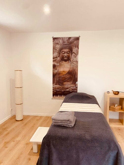 Une salle de massage sereine avec un lit simple recouvert d'une couverture gris foncé et deux serviettes pliées. Une grande tapisserie de Bouddha est accrochée au mur blanc. Un parquet en bois et une étagère en bois minimaliste sur laquelle se trouve un bol accentuent l'espace, créant une atmosphère tranquille. Une grande lampe se trouve dans le coin. - Bravadenn