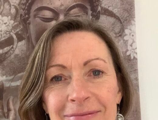 Une femme aux cheveux châtains courts sourit légèrement devant une grande tapisserie détaillée de Bouddha. Elle porte un haut gris clair et des boucles d'oreilles pendantes. Les motifs complexes reflètent la sérénité que l'on retrouve dans les pratiques de réflexologie plantaire souvent célébrées à Ploumoguer. - Bravadenn
