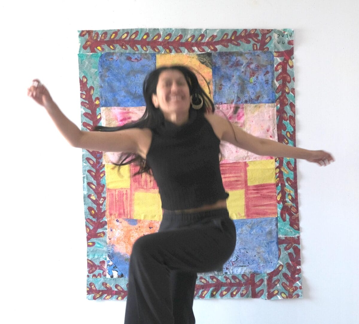 Une femme vêtue d'un haut noir sans manches et d'un pantalon noir saute énergiquement devant un patchwork coloré accroché à un mur blanc. Sa pose est dynamique et floue, masquant les traits de son visage, et ses cheveux sont libres et flottants. - Bravadenn