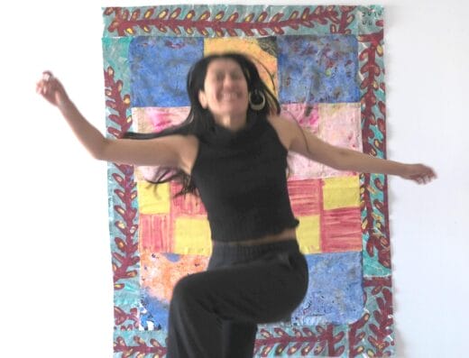 Une femme vêtue d'un haut noir sans manches et d'un pantalon noir saute énergiquement devant un patchwork coloré accroché à un mur blanc. Sa pose est dynamique et floue, masquant les traits de son visage, et ses cheveux sont libres et flottants. - Bravadenn