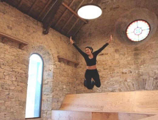 Une femme en tenue de sport noire saute les bras levés à l'intérieur d'une pièce rustique avec des murs en pierre, des planchers en bois, une fenêtre cintrée et un vitrail circulaire sous un haut plafond en bois avec des poutres apparentes. - Bravadenn